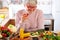 Vegetarian lifestyle. Beautiful elderly woman in the kitchen with a red wineglass in hand, looks on the table mix the of raw
