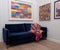 Velvet blue sofa with colourful wool blanket throw, in minimalist setting with white walls and colourful artwork.