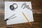 A vernier caliper holding a bearing, a pencil and a pair of compasses lying over drafting paper on wood background.