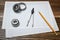 A vernier caliper holding a bearing, a pencil and a pair of compasses lying over drafting paper on wood background.