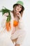 Vertical beauteous pregnant woman green hat, orange top and flax clothes, naked belly looking camera, orange carrots.