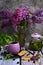 Vertical composition of a cup of herbal tea, a teapot, cookies and a bouquet of lilacs in a glass vase on a dark