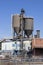 Vertical exterior of a brick and metal industrial building with storage silos, a dust collection system, and a conveyor belt