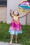 Vertical frontal photo of little girl in rainbow coloured summer dress throwing multicolored beach ball