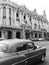 Vertical grayscale shot of a retro car driving in the street next to the Great Theatre of Havana