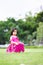 Vertical image. Asian baby child girl sitting in the green grass. Upset sad face kid. Children wearing dark pink dress.