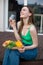 Vertical laughing brunette woman in green top and jeans drink wine and eating tasty food on plate, sit on wooden floor