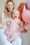 Vertical laughing and fooling blonde woman, mother holding baby in pink dress and flower bandelet, celebrating one year