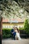 The vertical photo of the sensual newlyweds sitting on the bench under the blooming white tree. The groom is kissing the