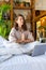 Vertical photo of a woman yoga practicing in bed in front of laptop monitor.