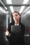 Vertical photo of young calm businesswoman holding laptop in her hands and looking at the camera in the lift. Female employee