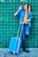 Vertical photo of a young girl waving to another person leaning against a green wall with a blue rolling suitcase