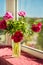 Vertical picture of bouquet of maroon and pink peonies in glass vase on windowsill in sunny day. Image with selective focus.