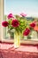 Vertical picture of bouquet of maroon and pink peonies in glass vase on windowsill in sunny day. Spring summer floral background