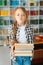 Vertical portrait of joyful elementary child school girl holding stack of books in library at school, looking at camera.