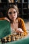 Vertical portrait of serious young woman in elegant eyeglasses making chess move with knight piece sitting in floor