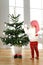 Vertical portrait of small blonde female child standes bare foot on wooden floor, decorates New Year tree, wears Santa s