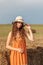 Vertical portrait of a young cute rural woman in an orange sarafan and a hat near a stack of wheat hay on a field in the rays of t