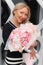 Vertical positive smiling mature blond businesswoman in striped dress carry amazing pink flower bouquet. Corporate party