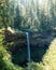 Vertical shor of a narrow little waterfall flowing over the clifs in the dense pine forest