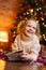 Vertical shot of cheerful dreaming little blonde child girl writing letter to Santa Claus lying on floor on background