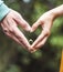 Vertical shot of a heart of hands by a couple holding an engagement ring on a greenery background