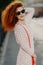 Vertical shot of lovely woman in trendy sunglasses, has curly bushy red hair, carries small bag, poses outdoor over blurred