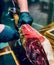 Vertical shot of a person in black plastic gloves slicing a Spanish Jamon leg