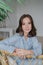 Vertical shot of pleasant looking young European dark haired woman sits on wicker chair, dressed in striped jacket, has red manicu