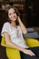 Vertical shot of pretty girl dressed in white t shirt and jeans, has happy look at camera, sits at yellow armchair, being in good