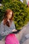 Vertical shot of redhead young woman standing in city park near trees holding laptop typing using outdoors Wi-Fi