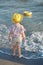 Vertical shot of small fashionable girl stands back, wears summer hat, blouse and shorts, admires sea, plays alone, breathes fresh