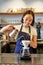 Vertical shot of smiling asian bartender, barista in blue apron, pouring water with small kettle, brewing coffee behind