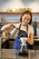Vertical shot of smiling asian bartender, barista in blue apron, pouring water with small kettle, brewing coffee behind