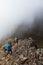 Vertical shot of two hikers peeking out of the edge of a ravine on a rocky mountain with clouds of mist on top of Cerro Chirripo