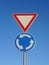 A vertical shot of two traffic signs on the same post - Give way and blue roundabout signs