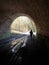 Vertical shot of an unrecognizable person with a dog going out of a tunnel with a muddy road