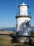 Vertical shot of a white lighthouse near the seashore under the shadow of a tree on a sunny day