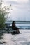 Vertical smiling barefoot romantic overweight plump multiracial dark skin woman sit by sea or river coast near bridge