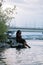 Vertical smiling serene barefoot curly plump Afro-American multiracial woman sit by the river coast. Tranquil vacation