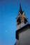 Vertical of the spire of the San Vigilio church with clock and moon in the sky.