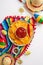 Vertical top view of a sombrero, poncho, maracas, tequila shots, lime wedges, chili peppers, nacho chips, and salsa arranged