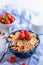 Vertical trendy cereal pancakes in blue bowl with strawberries and hazelnuts on blue gauze on white wooden background