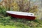 Very small plastic white and red river boat left on side of river bank surrounded with grass and dense branches