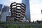 Vessel  TKA, a spiral endless staircase, with people walking around, skyscrappers behind, flowers in front of it, Hudson Yards