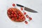 Vibrant small red tomatoes with green vines on wooden chopping board, large chef knife near, white stone table under, view from