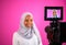 Videographer in pink studio recording video on professional camera by shooting female muslim woman