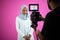 Videographer in pink studio recording video on professional camera by shooting female muslim woman