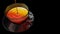 View from above of a jet of liquid filling a glass cup full of tea on glass dish on black background.