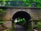 View of alley going through mossy stone arch bridge with metal fence above surrounded by greenery